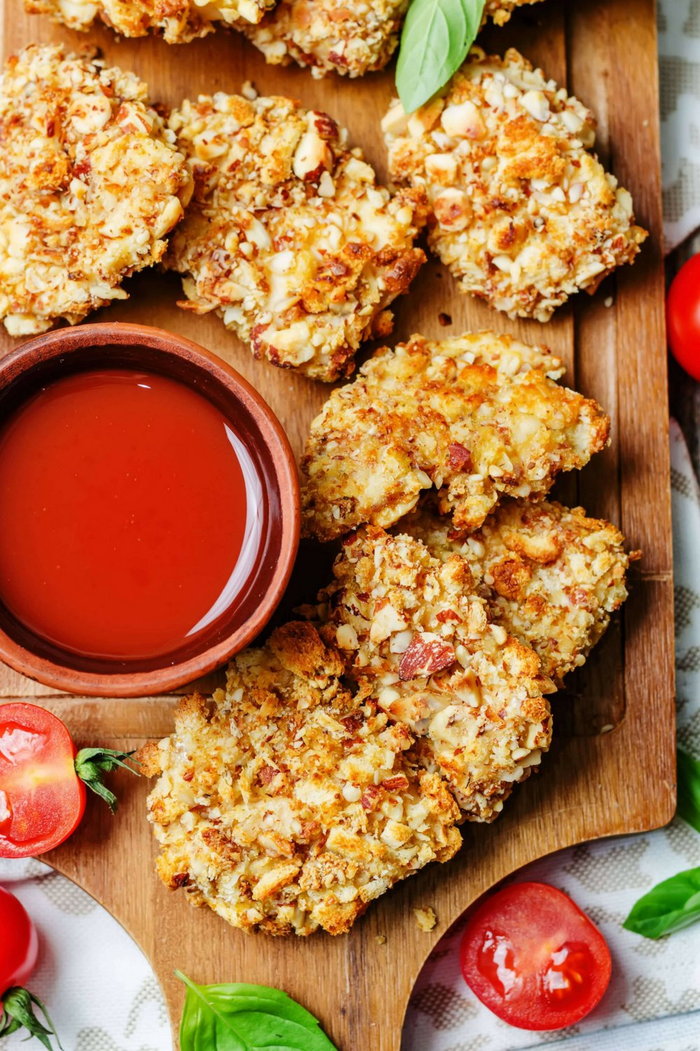Low carb almond crusted chicken tenders on a wood cutting board with tomatoes and ketchup.