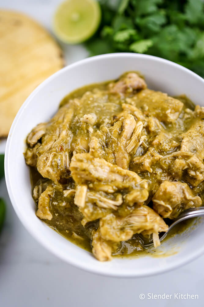 Chile verde recipe in a white bowl with tortillas, cilantro, and limes.
