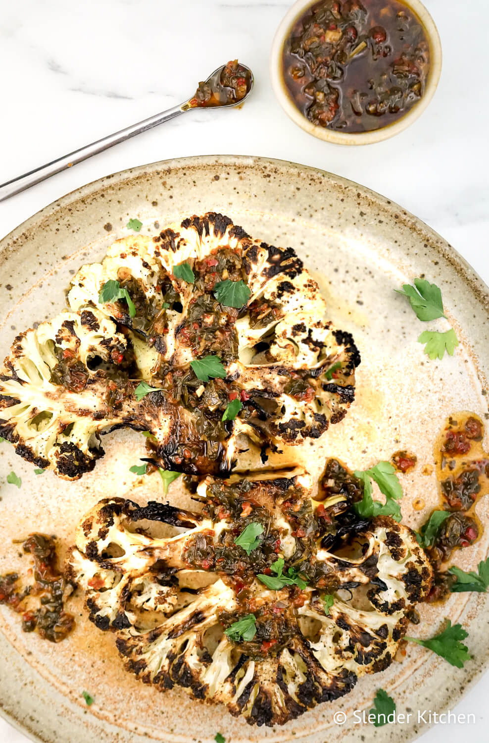Roasted cauliflower steaks on a clay plate with chimichurri on top and a marble white background.