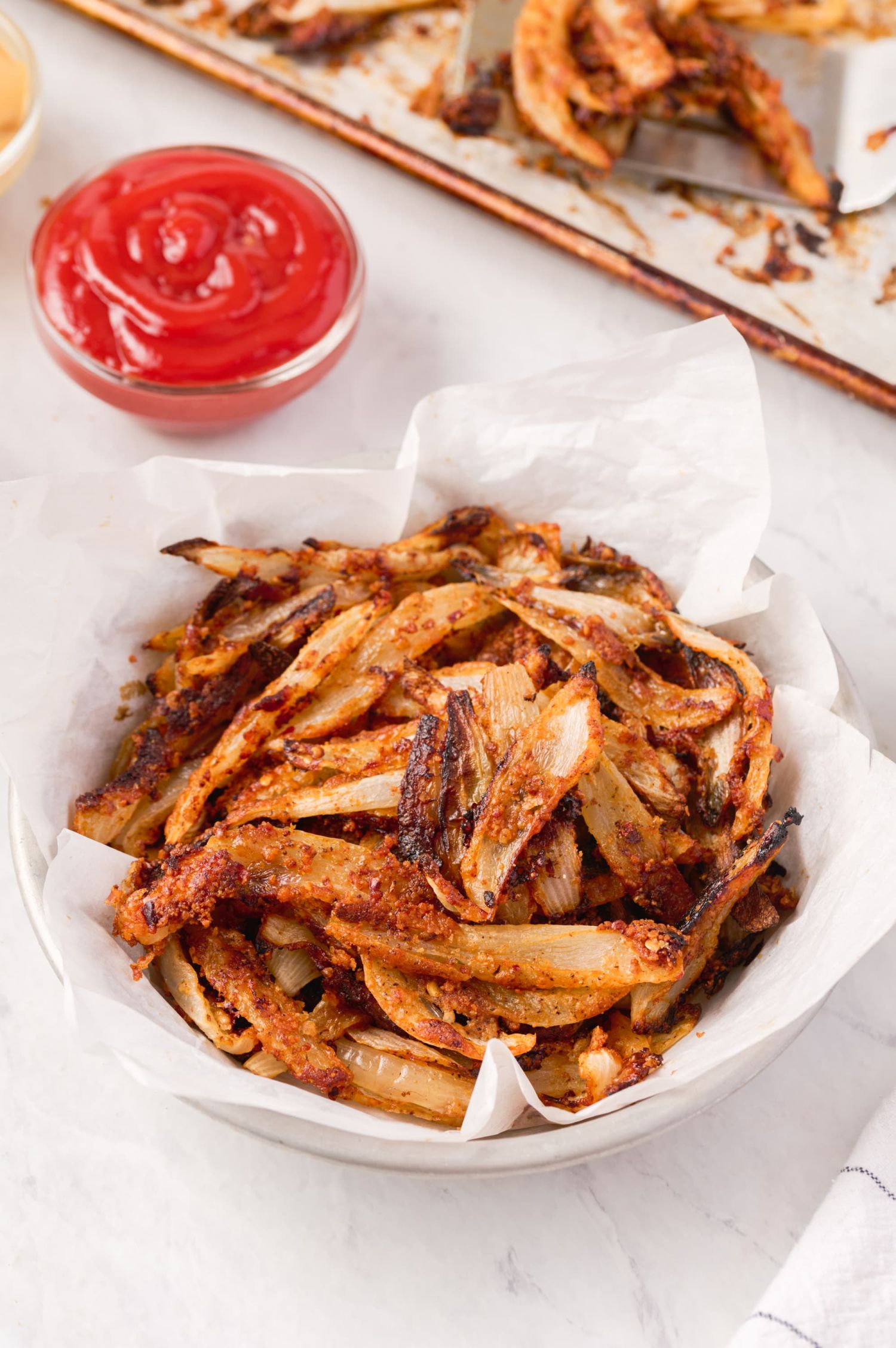 Onion straws that have been baked with a crispy Parmesan coating in a white bowl with ketchup on the side.