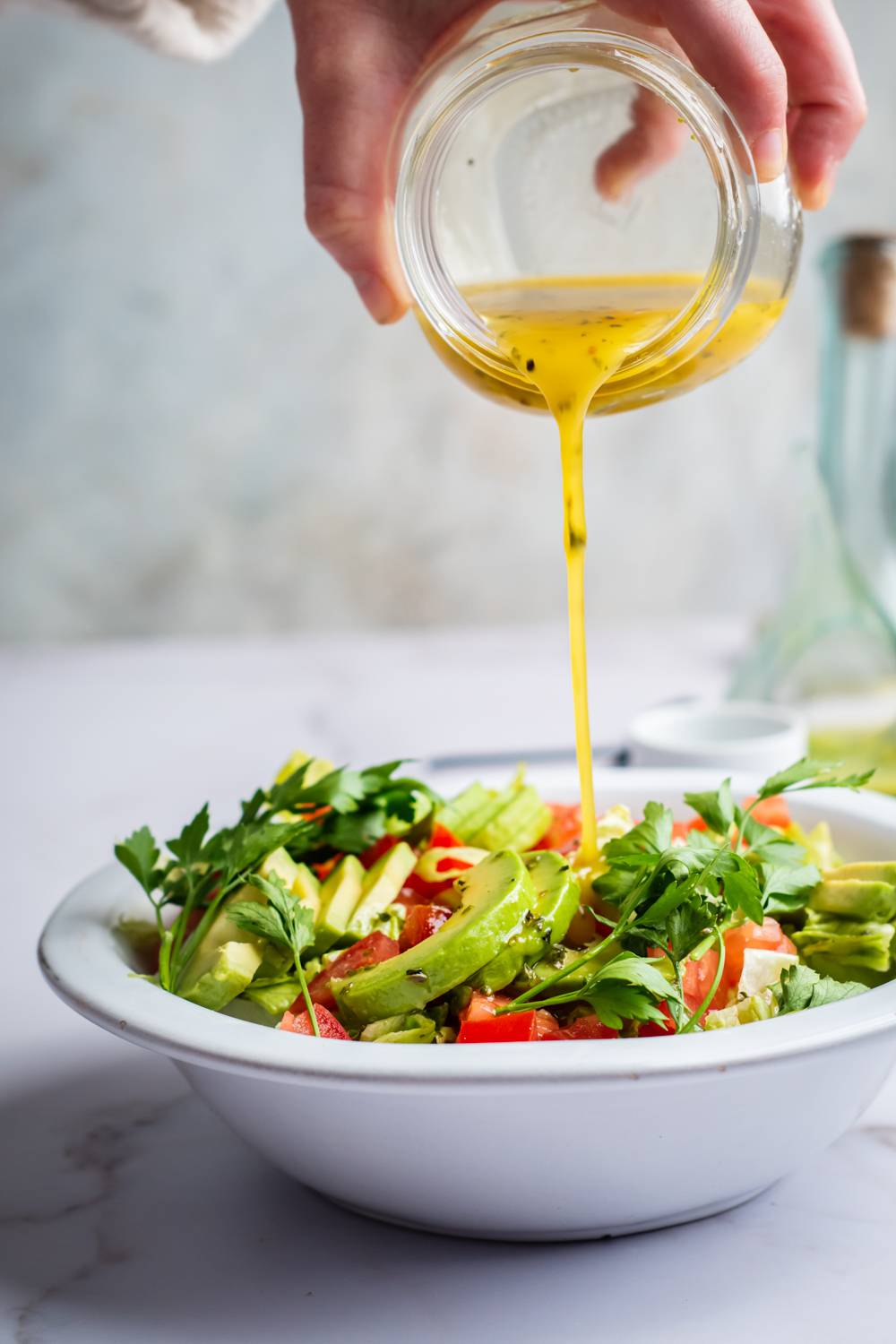 Italian dressing being poured over a salad with avocado, tomatoes, and herbs.