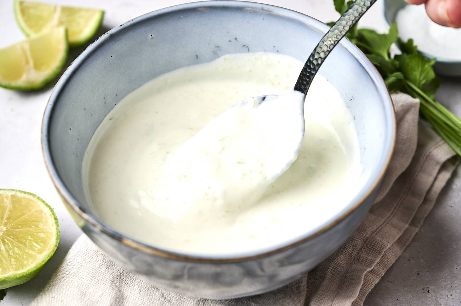 A light green creamy sauce in a blue bowl, with a spoonful lifted, surrounded by fresh lime slices and cilantro.