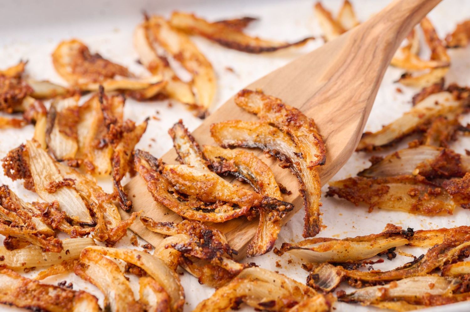 Onion straws coated with Parmesan cheese and spices on a baking sheet with a saptula.