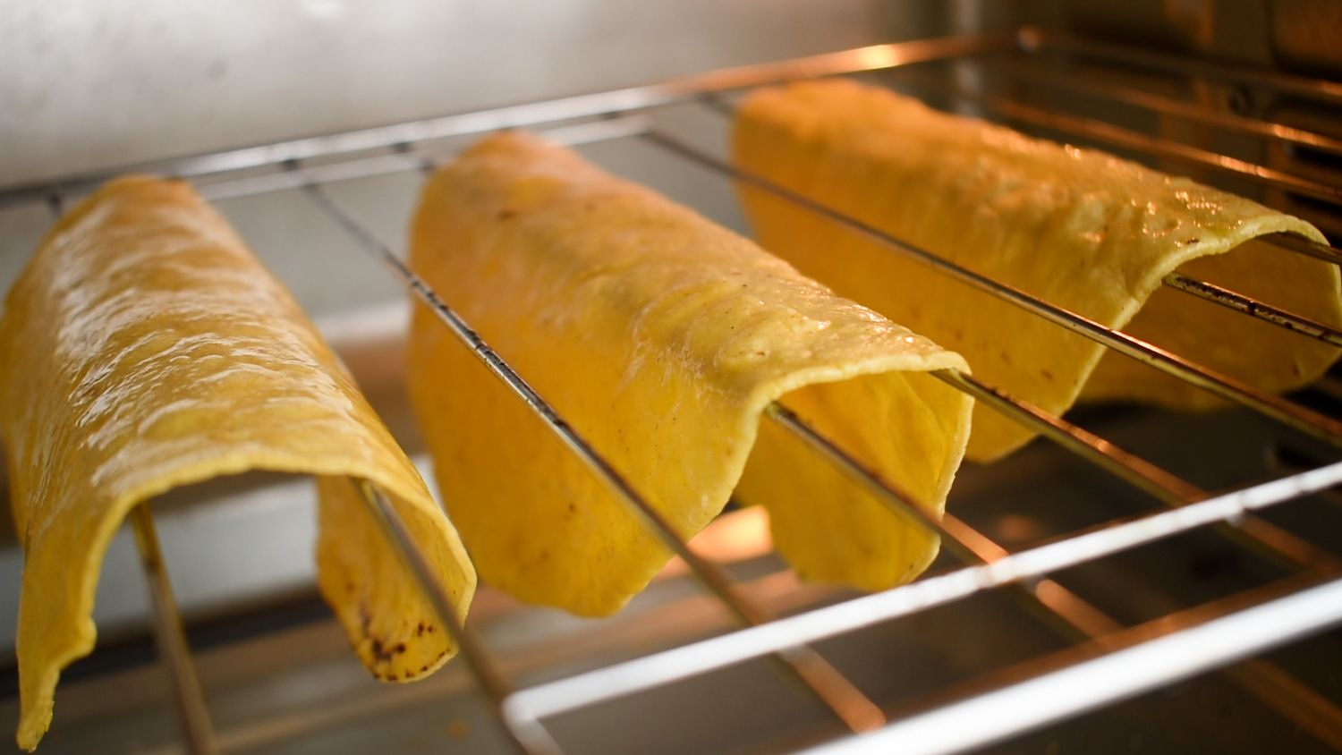 Corn tortillas baking in the oven to make taco shells that are draped over the oven rack.