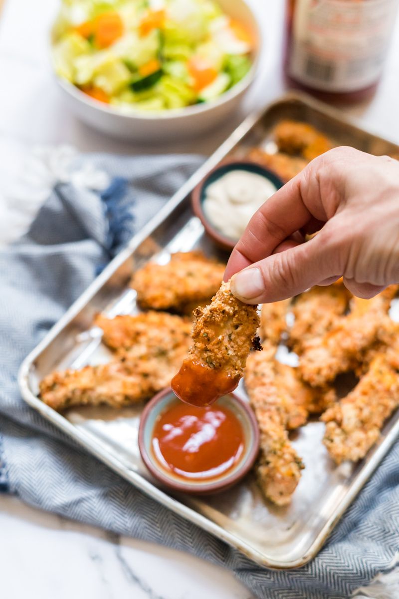 Crispy buffalo ranch chicken tenders being dipped in buffalo sauce with salad on the side.
