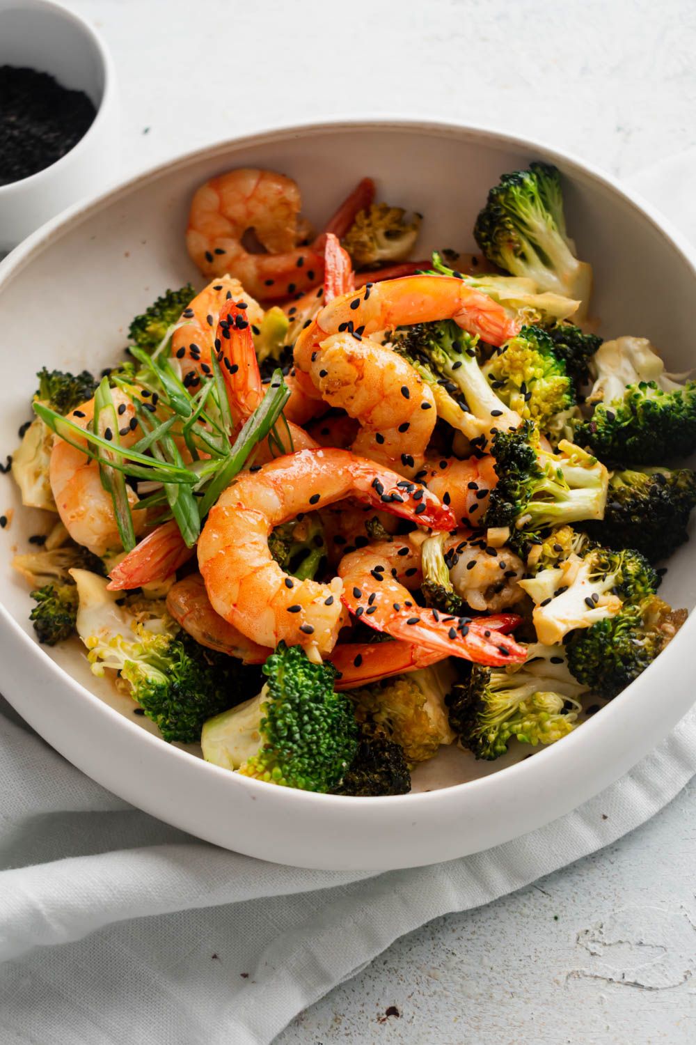 Firecracker shrimp in a slightly spicy sauce served with broccoli, green onions, and black sesame seeds in a white bowl.