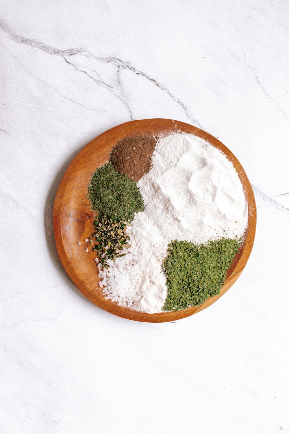 Ingredients for ranch seasoning with buttermilk powder, dried herbs, onion powder, garlic powder, and pepper on a wooden plate.
