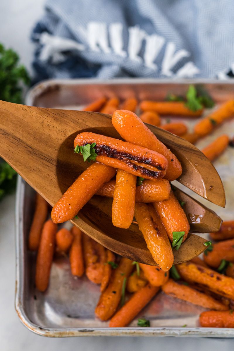 Roasted baby carrots with olive oil, honey, salt, and pepper served on a baking sheet with a wooden spoon and parsley.