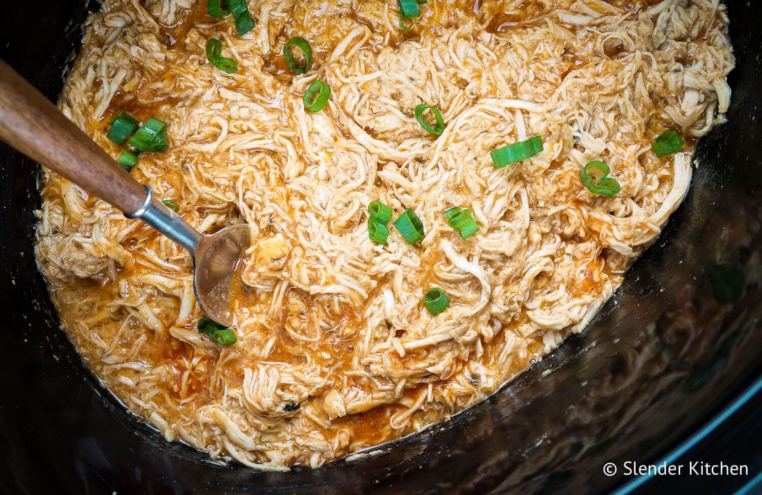 Crockpot buffalo chicken shredded with buffalo sauce and green onions in a black slow cooker.