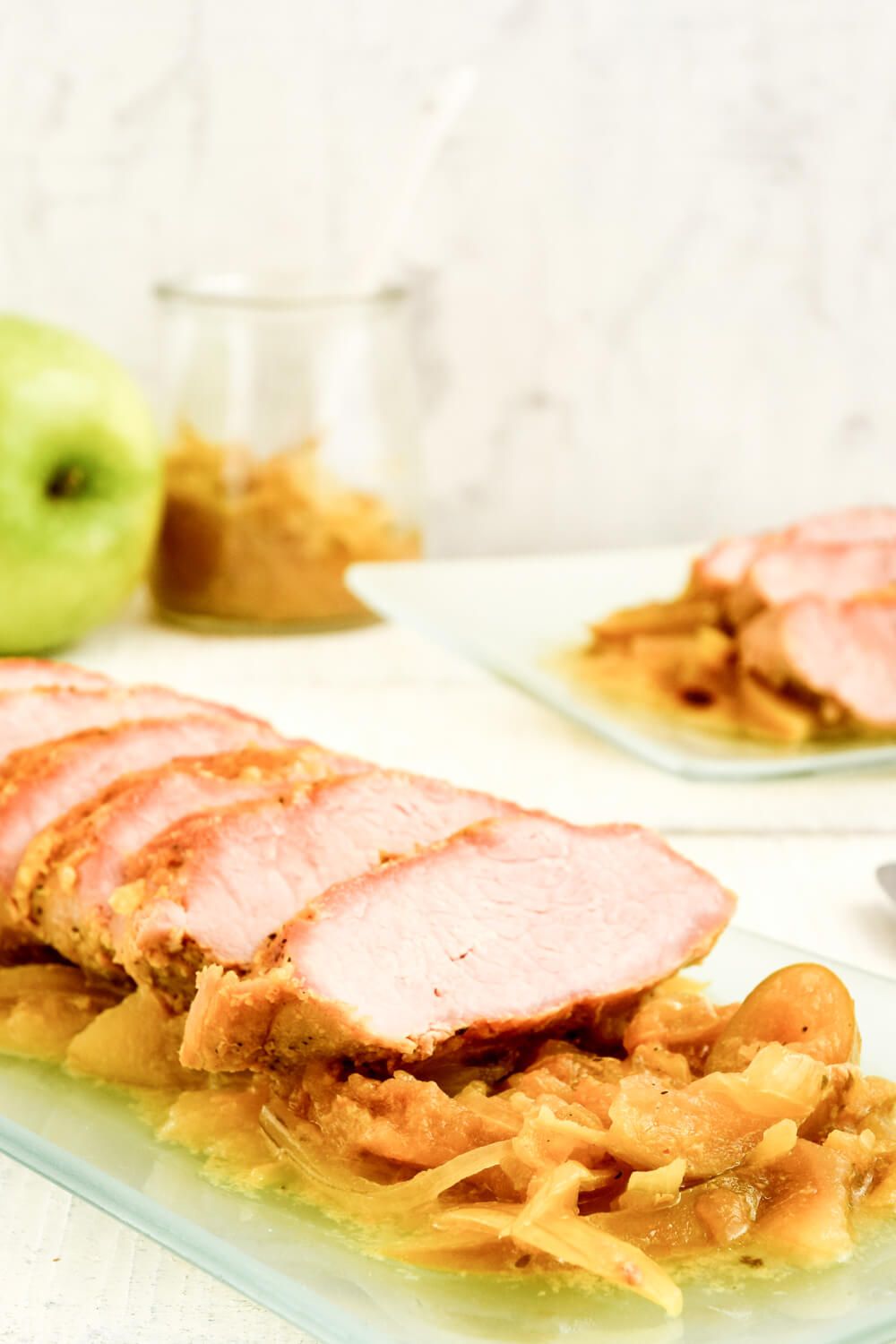 Crockpot pork tenderloin with apples on a glass plate with an apple and applesauce in background.