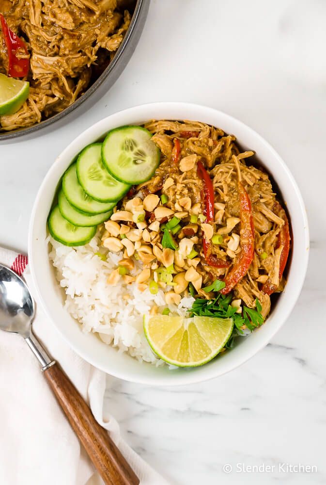 Thai peanut chicken made in the slow cooker in a bowl with rice, red peppers, peanuts, cilantro, and limes.