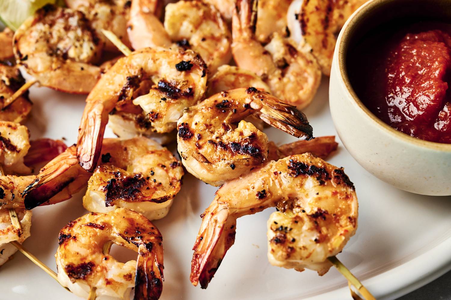 Grilled shrimp skewers arranged on a white platter, served with lime wedges and a bowl of red dipping sauce.