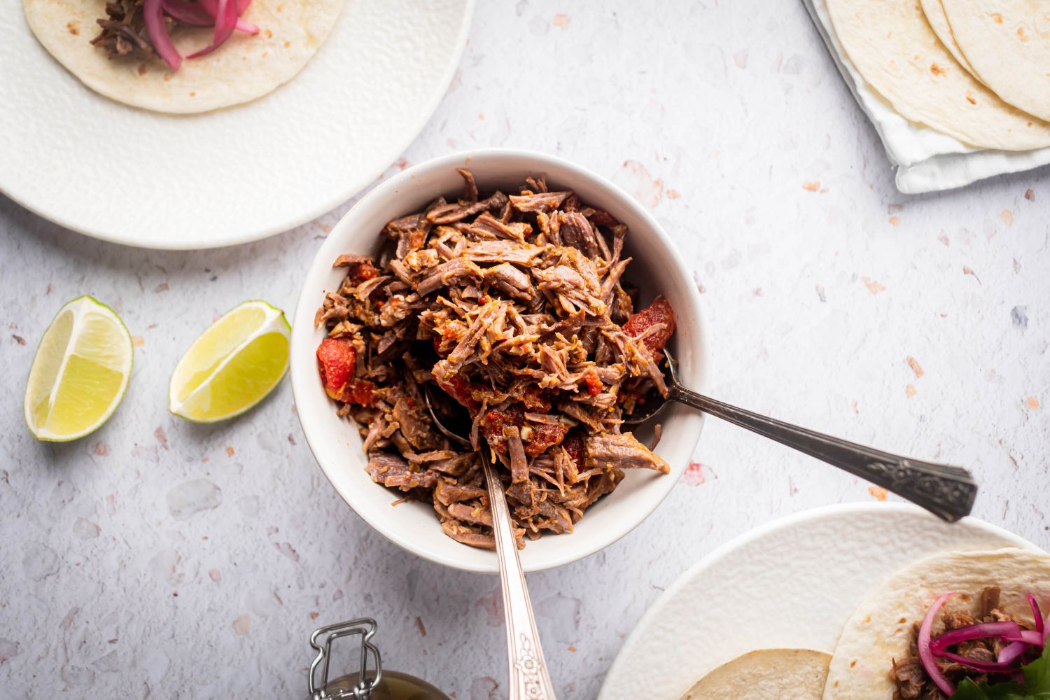 Mexican shredded beef in a bowl with tomatoes and chipotle peppers with tacos on the side.