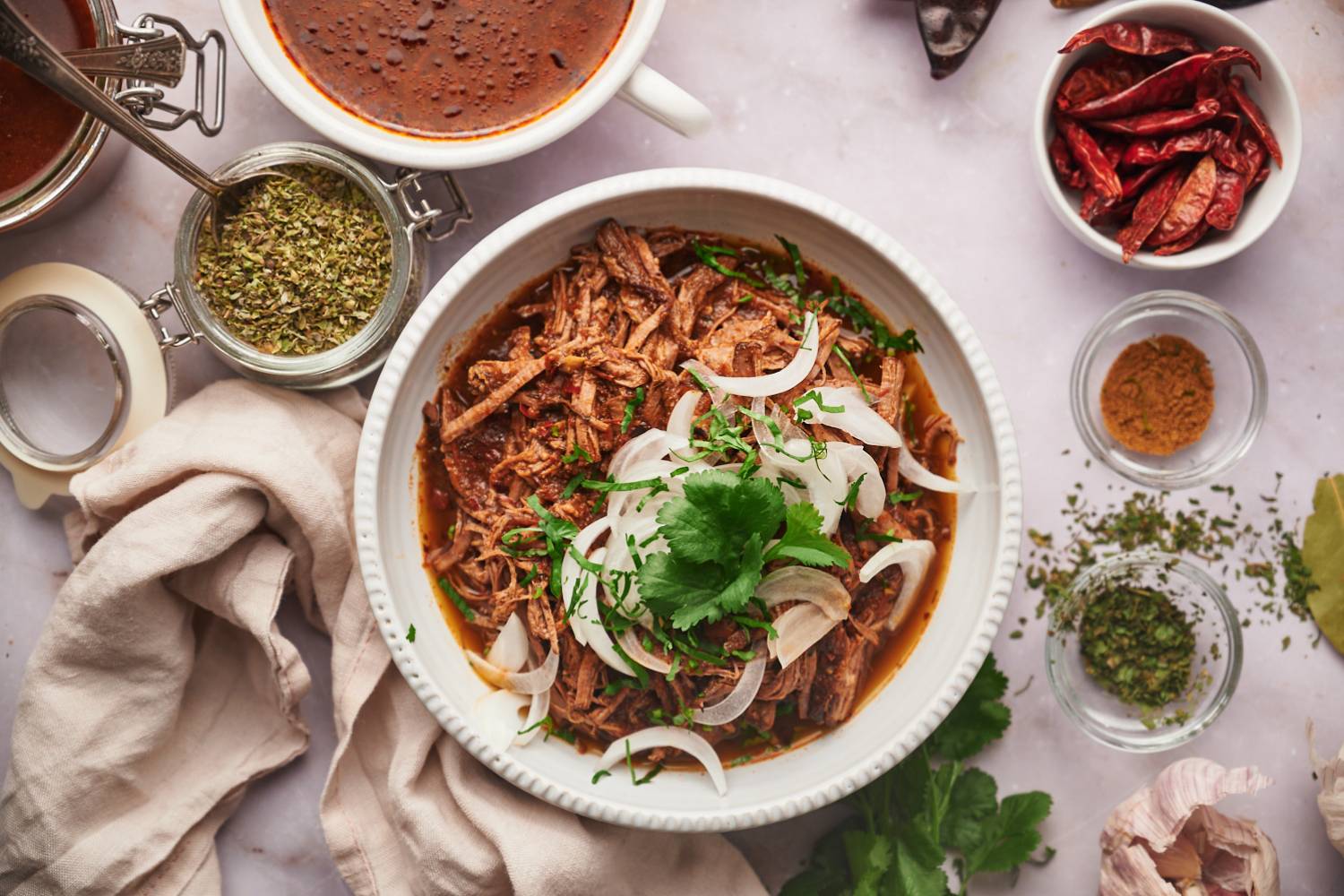 Birria de res with a red chile sauce served in a bowl with corn tortillas, sliced onions, cilantro, and consome.