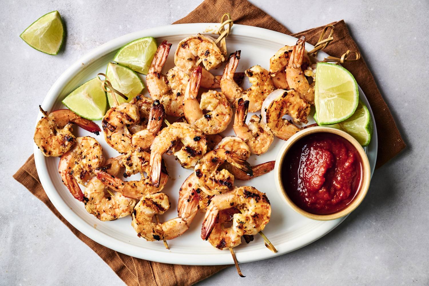 Spicy Grilled Shrimp Grilled shrimp skewers served with lime wedges and a bowl of dipping sauce on a white platter over a brown cloth.