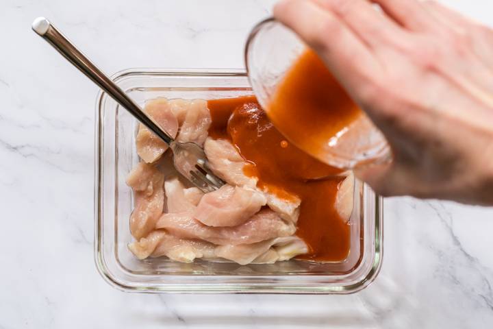 Chicken strips being marinated in a glass container with buffalo sauce.