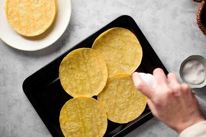 Corn tortillas on a baking sheet being sprayed with cooking spray.