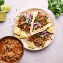 Slow cooker beef barbacoa tacos on corn tortillas with red onion, cilantro, and lime wedges.