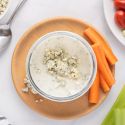 Yogurt blue cheese dressing with crumbled blue cheese in a glass bowl with raw vegetables on the side.