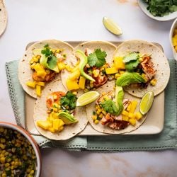 Salmon tacos with corn salsa, mango, fresh avocado slices, and lime wedges on a tray. 