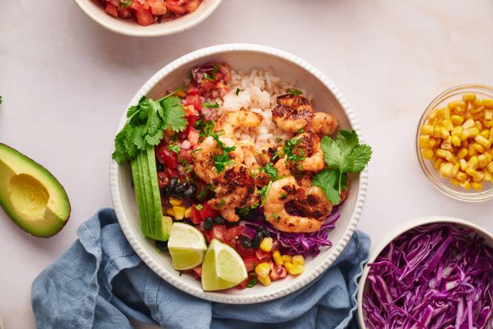 Shrimp taco bowls with cooked shrimp, avocado, rice, lime, cilantro, cabbage, corn, and pico de gallo in a white bowl.