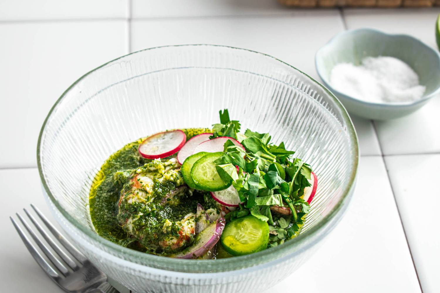 A vibrant bowl of green sauce with shrimp, garnished with radishes, cucumber, and fresh herbs, beside a small bowl of salt.
