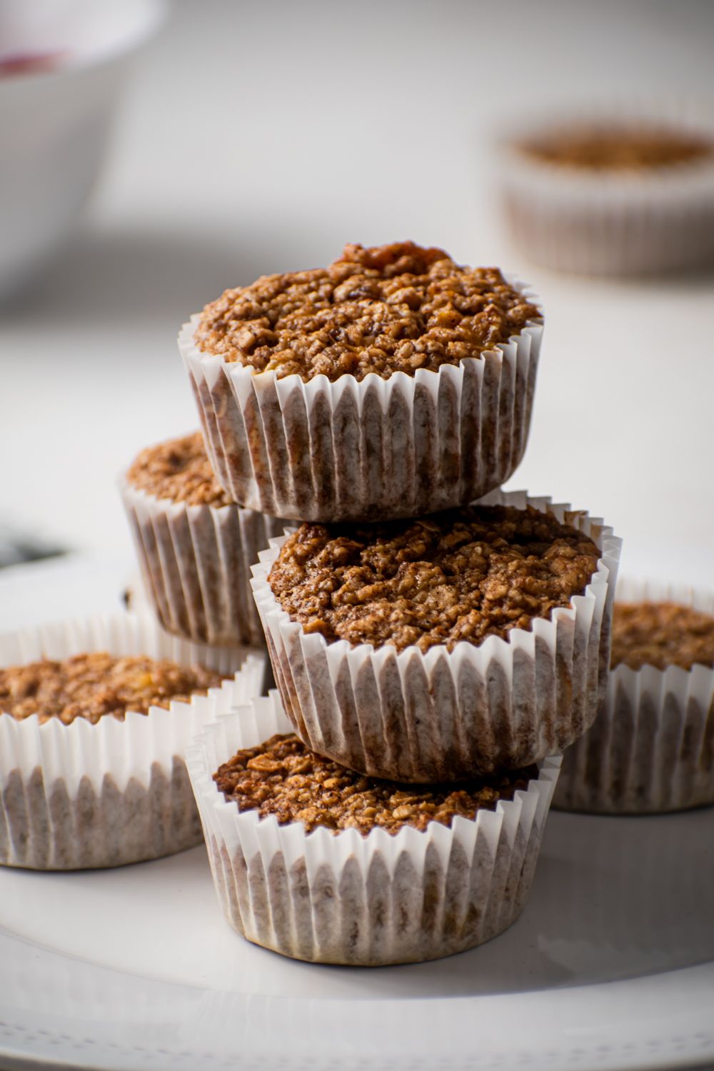 Baked oat muffins with white liners stacked on a plate with rolled oats, bananas, and almond milk.