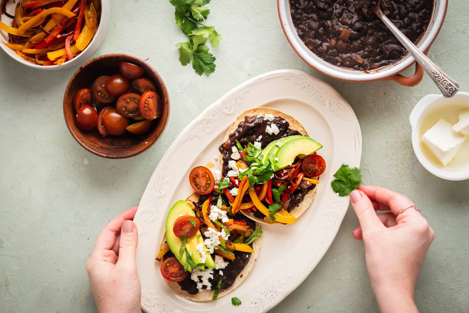 Two black bean tostadas topped with sliced avocado, colorful bell peppers, cherry tomatoes, crumbled cheese, and fresh cilantro on a white oval plate. Surrounding bowls contain cherry tomatoes, sautéed peppers, black beans, and cheese.