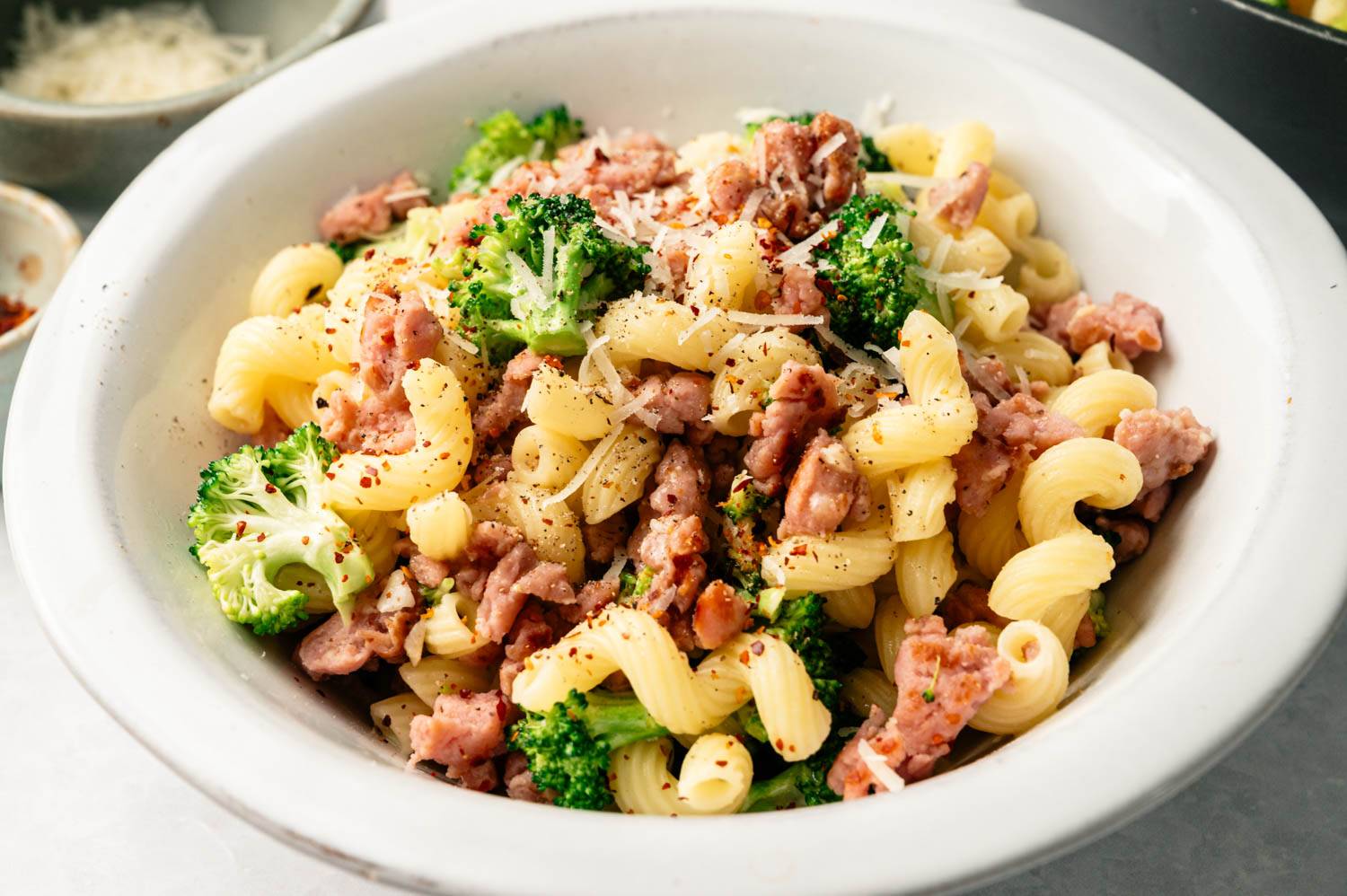 A bowl of corkscrew pasta with broccoli and crumbled sausage, topped with grated cheese and pepper, creating a warm and hearty meal.