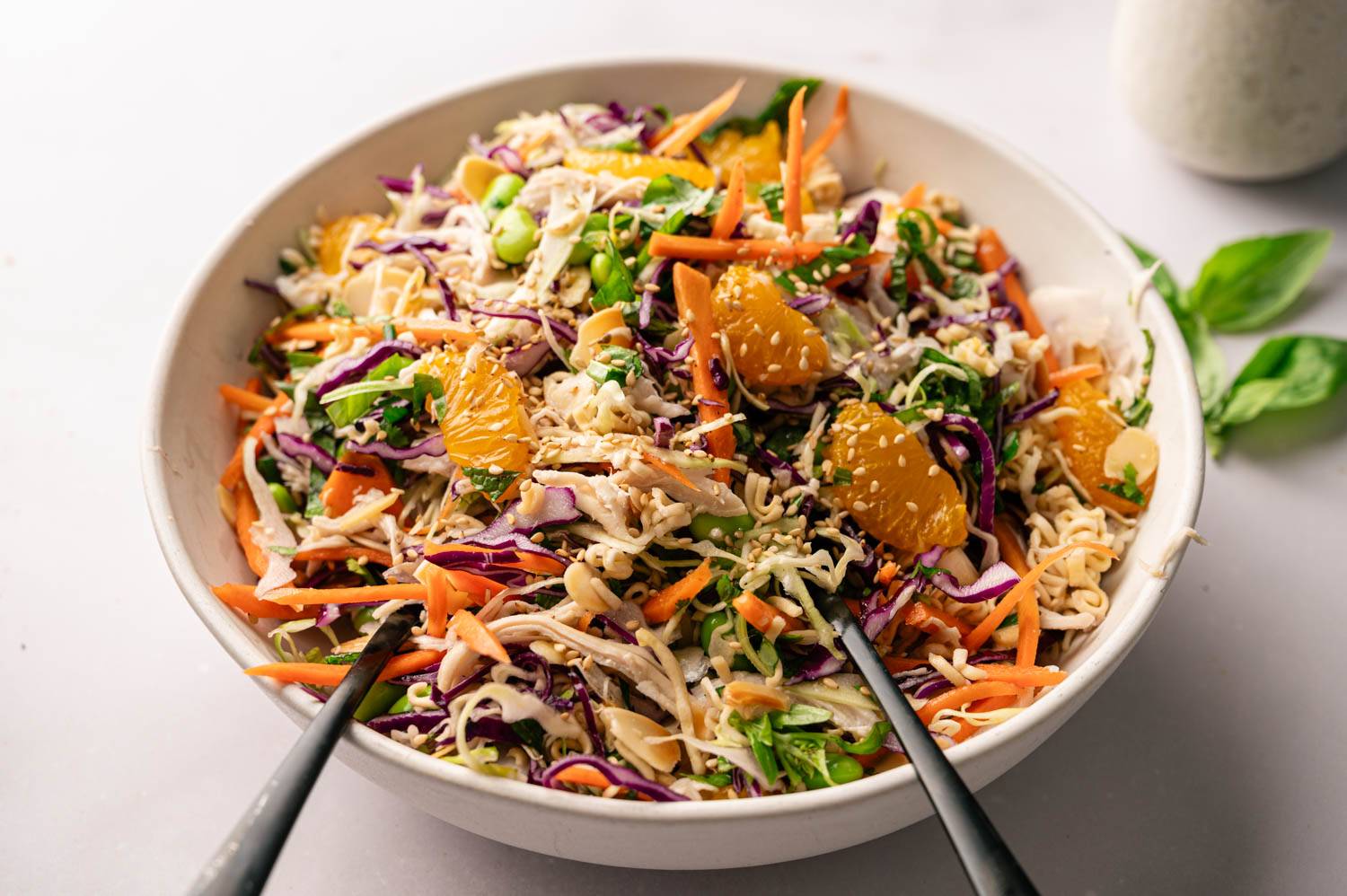 A vibrant Asian salad in a bowl with shredded carrots, cabbage, edamame, shredded chicken, orange segments, and sesame seeds, creating a fresh, colorful mix.