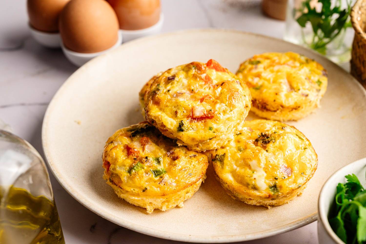 Plate of four golden-brown vegetable egg muffins, featuring visible herbs and vegetables, with eggs in the background. Cozy, inviting breakfast setting.