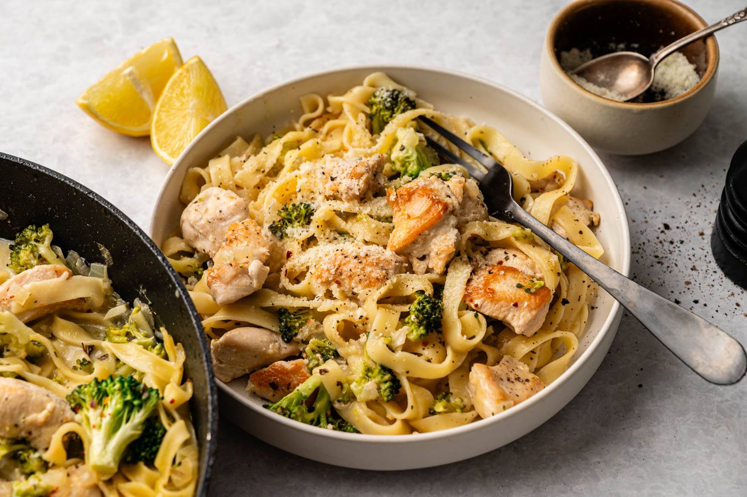 A bowl of creamy chicken fettuccine with broccoli and black pepper, garnished with Parmesan. Fresh lemon wedges are nearby, conveying warmth and comfort.