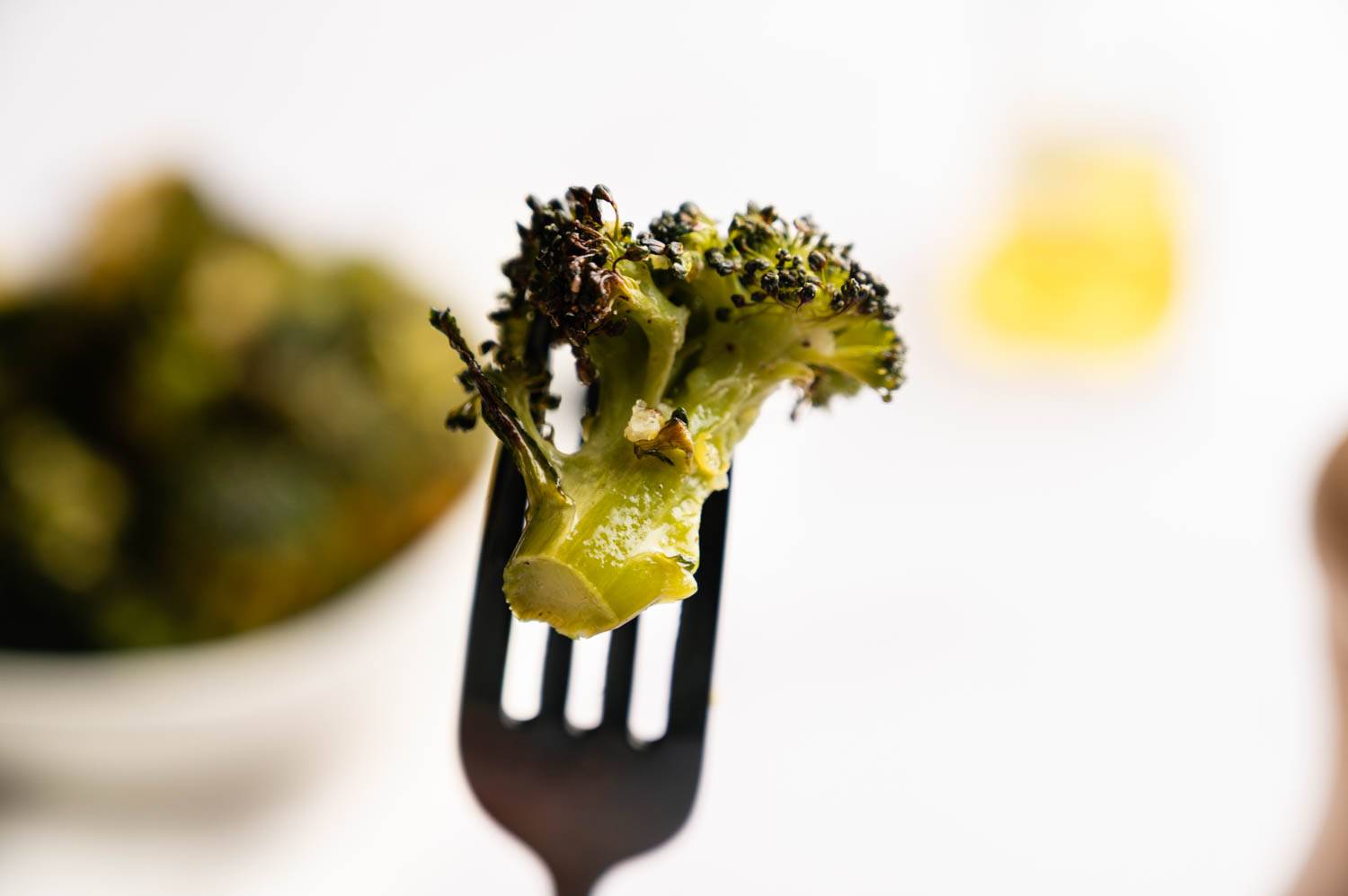 Close-up of a roasted broccoli floret on a fork, charred edges visible. Soft focus background with a bowl of broccoli and a blurred yellow spot.