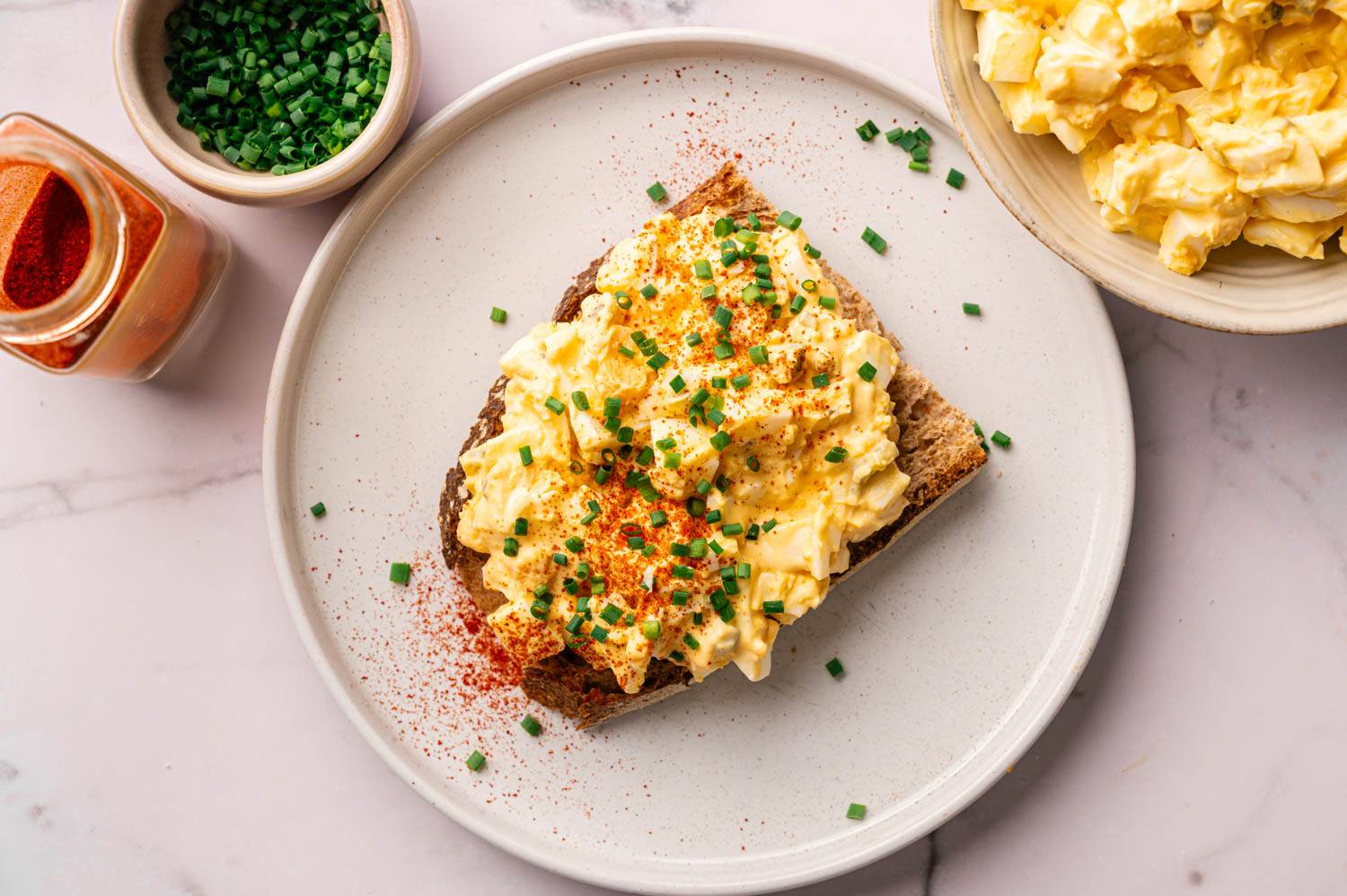 Open-faced egg salad sandwich garnished with chopped chives and paprika on a white plate, surrounded by bowls of chives, paprika, and extra egg salad.