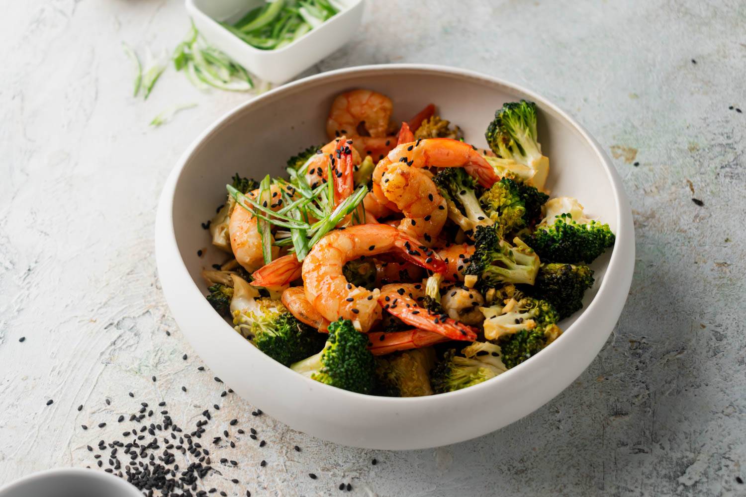 A bowl of firecracker shrimp and broccoli, garnished with sliced scallions and sesame seeds.