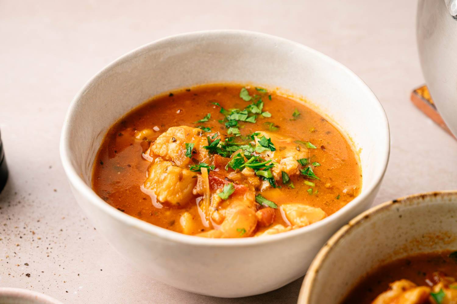 A white bowl of rich, orange seafood stew is garnished with fresh herbs. The soup appears hearty and flavorful, evoking warmth and comfort.
