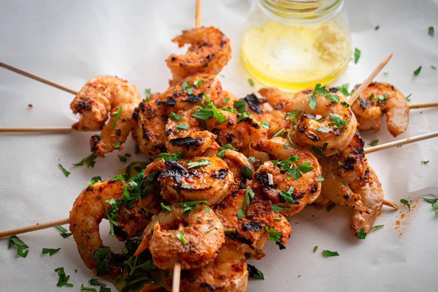 Grilled shrimp skewers garnished with parsley, served with a jar of olive oil on parchment paper.
