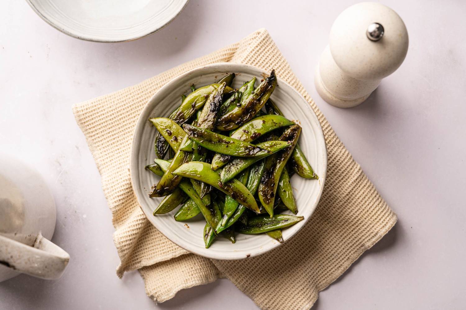 A white bowl filled with charred sugar snap peas on a beige cloth, next to a white pepper mill.