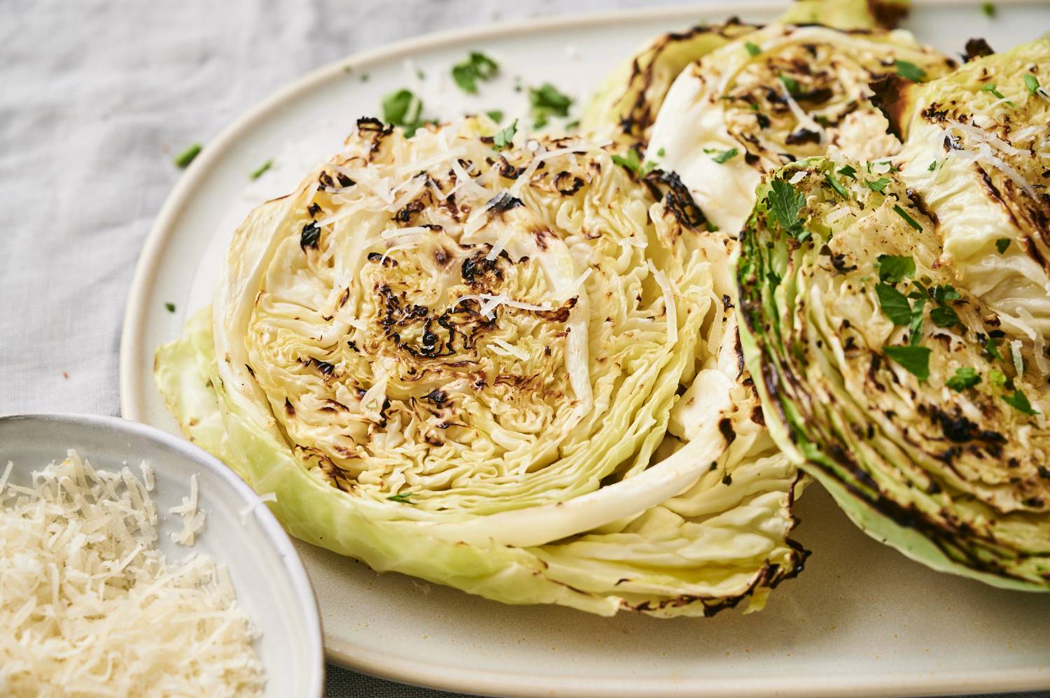 Grilled cabbage slices topped with shredded cheese and herbs, served on a light plate beside a bowl of extra cheese.