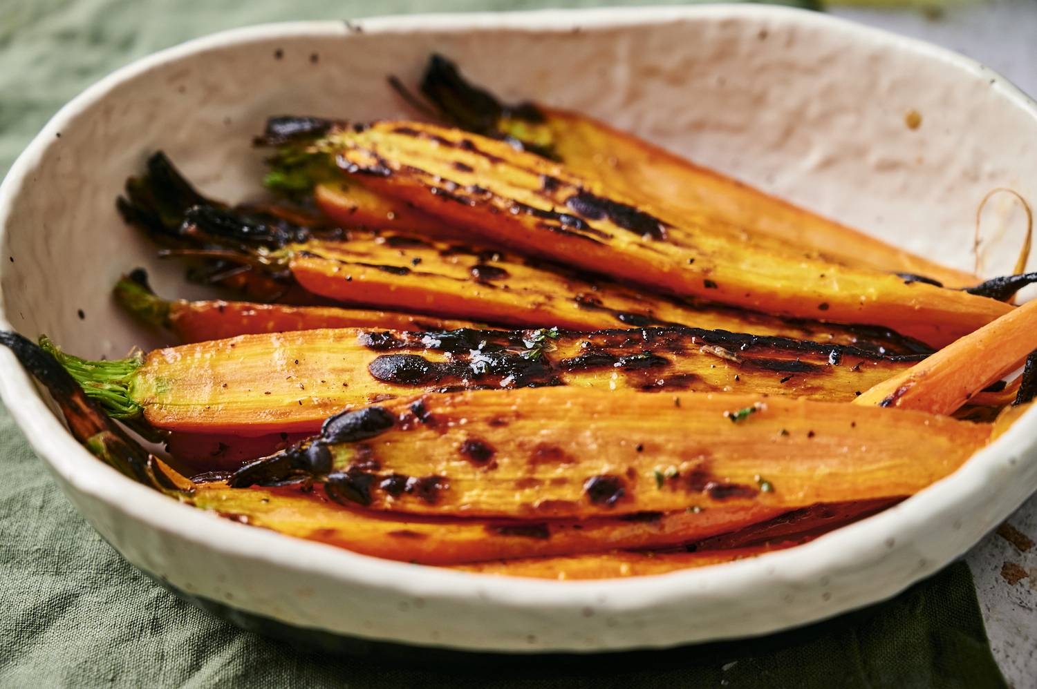 A white bowl filled with charred orange carrots, garnished with herbs, resting on a green cloth backdrop.
