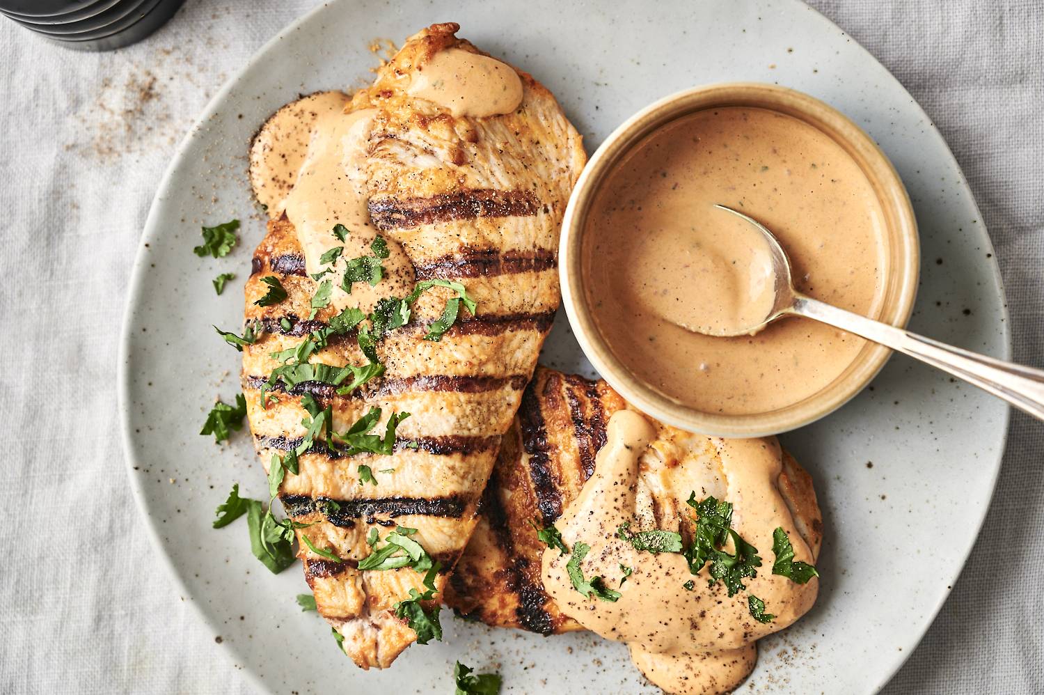 Grilled chicken breast with sauce and parsley on a plate beside a bowl of creamy dressing.