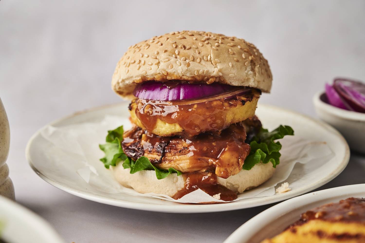A sesame seed bun sandwich stacked with lettuce, glazed chicken, grilled pineapple, and red onion, served on a plate.