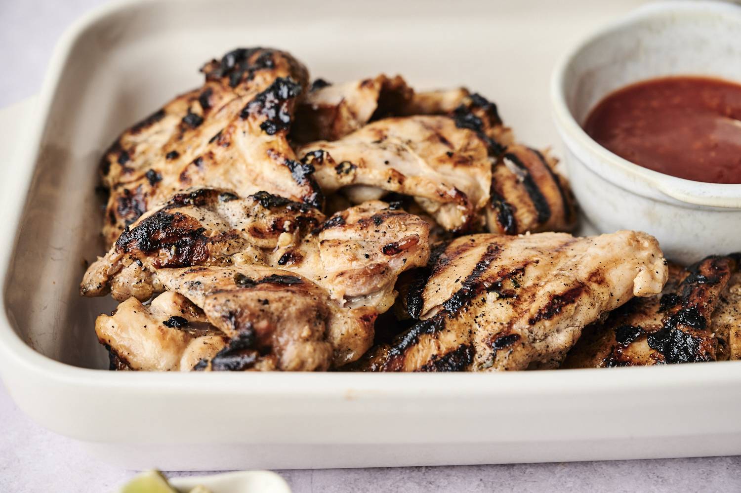 Grilled chicken thighs with char marks on a platter, accompanied by a small bowl of dipping sauce.
