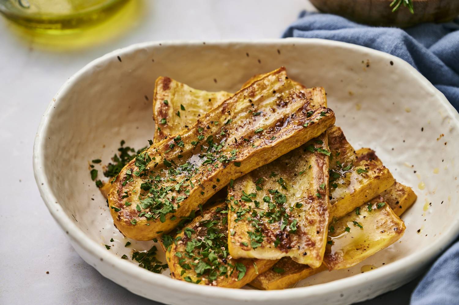 Golden, grilled squash slices garnished with fresh herbs, drizzled with oil, in a rustic white bowl, set against a soft neutral background.