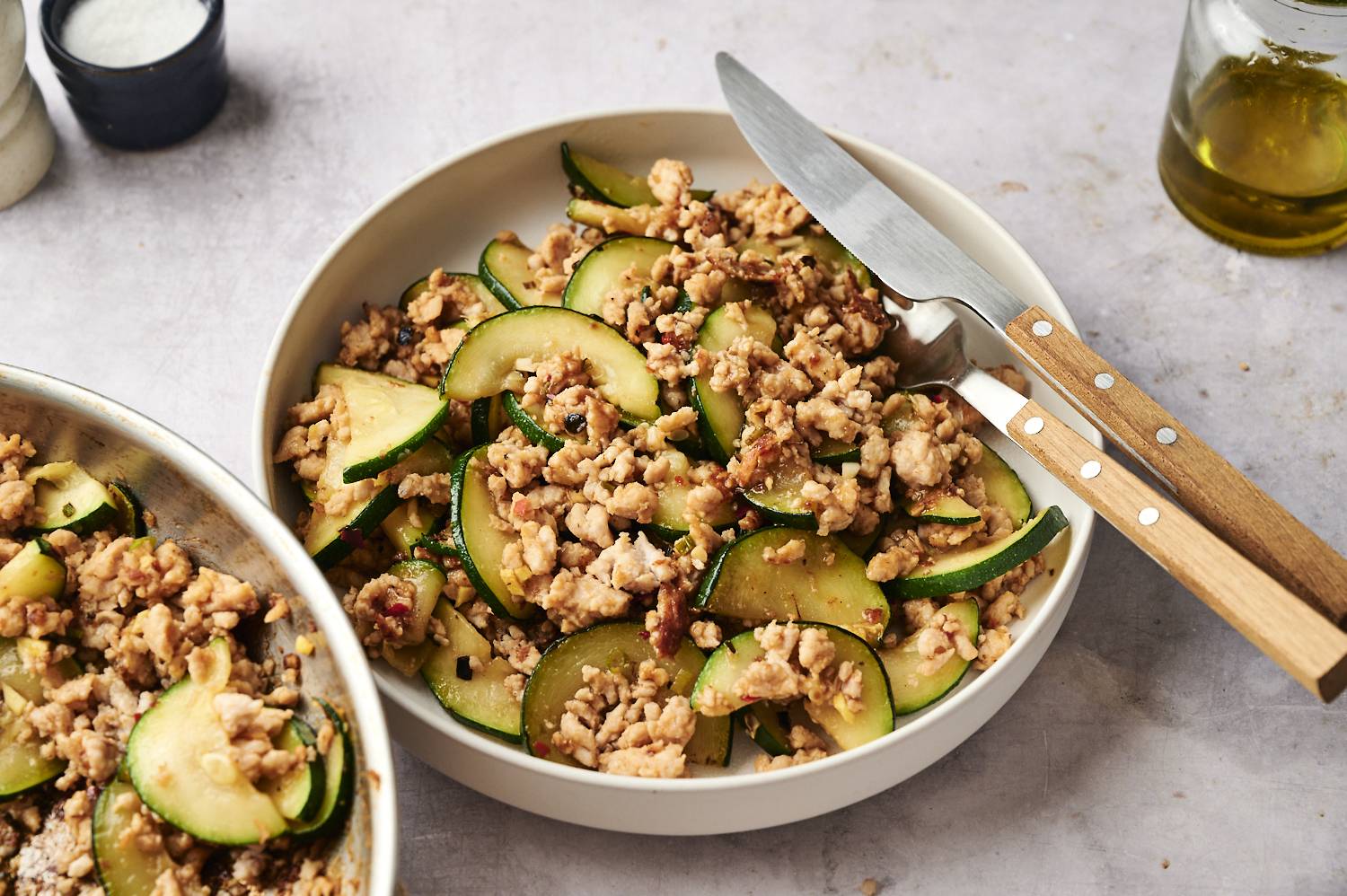 White bowl filled with ground turkey zucchini stir-fry, featuring browned ground turkey and sautéed zucchini slices, served with a knife and fork on the side.