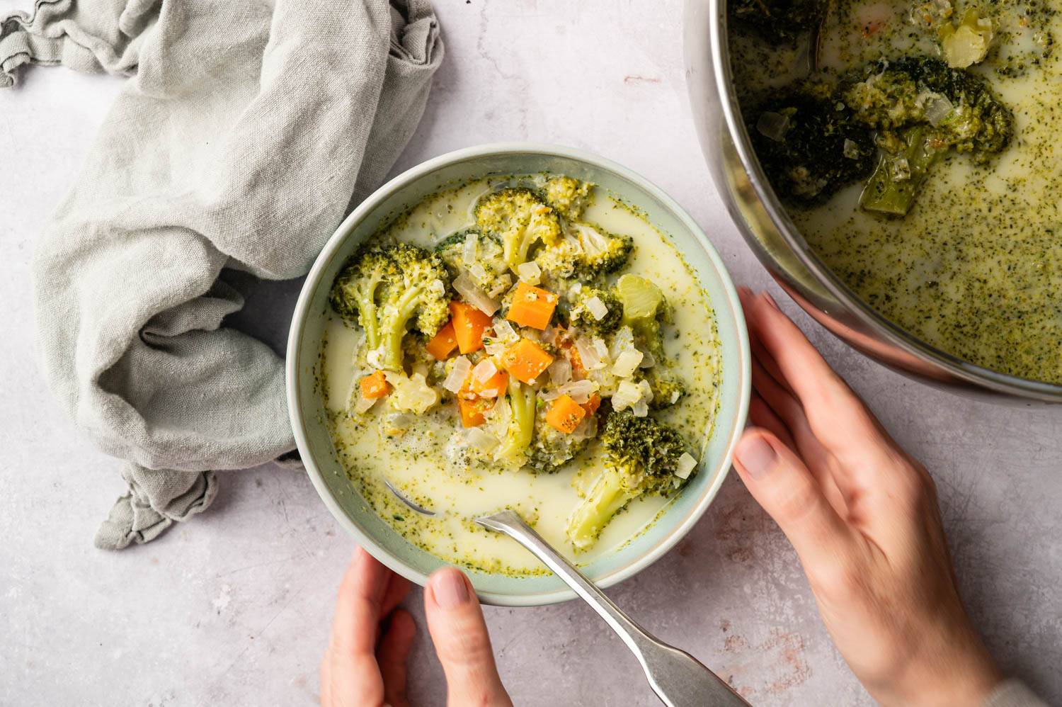 A bowl of creamy broccoli soup with carrots and onions, garnished with herbs. Hands hold the bowl near a pot. A soft, gray cloth is beside it.