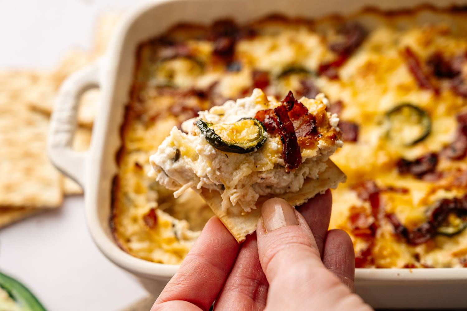 Hand holding a cracker topped with creamy, baked cheese, bacon, and jalapeño. Background shows a dish of the same cheesy, baked mixture.
