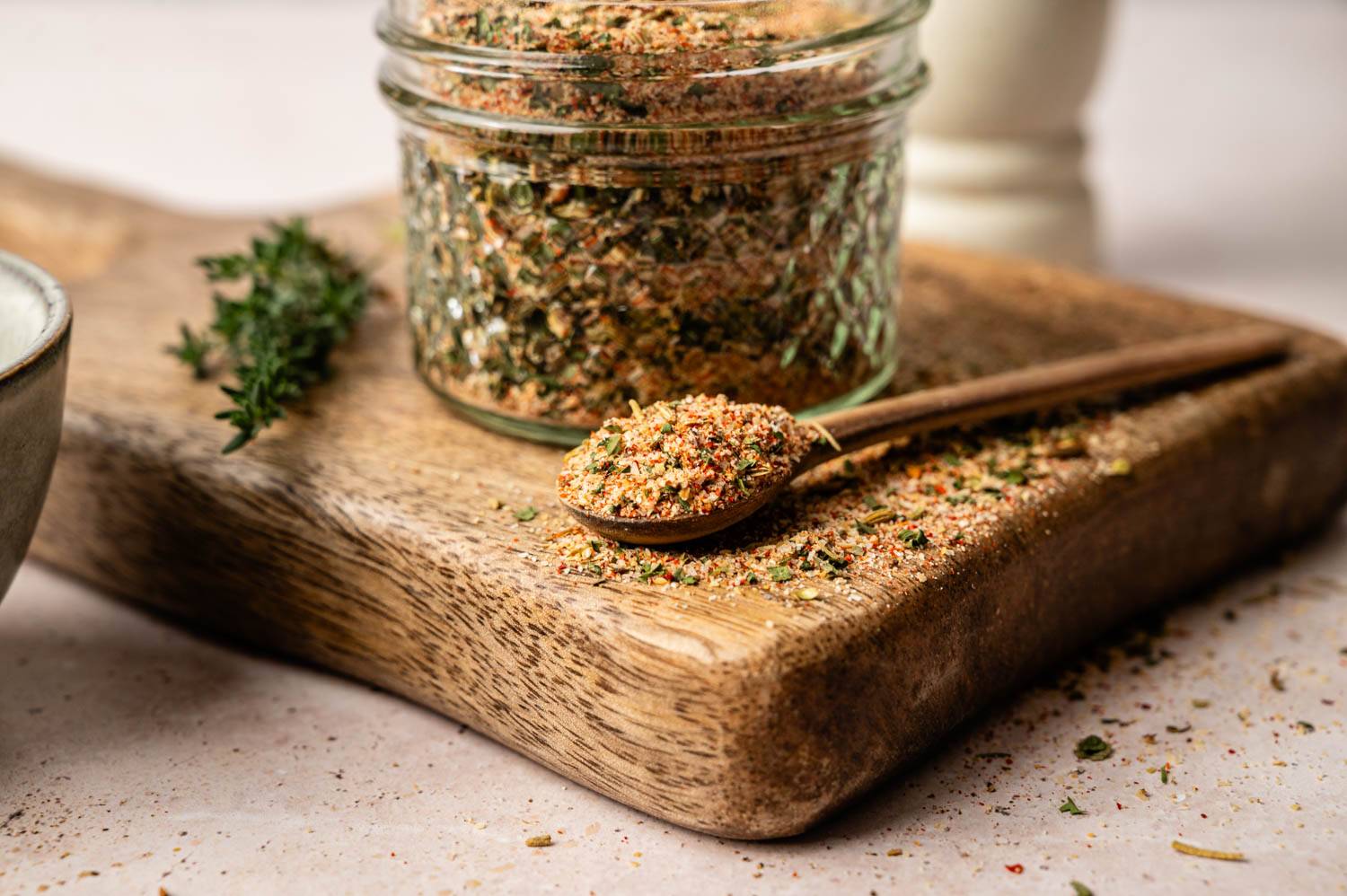 A rustic wooden board holds a jar of colorful seasoning blend, with a wooden spoon full of spices. Fresh thyme sprigs add a natural touch.