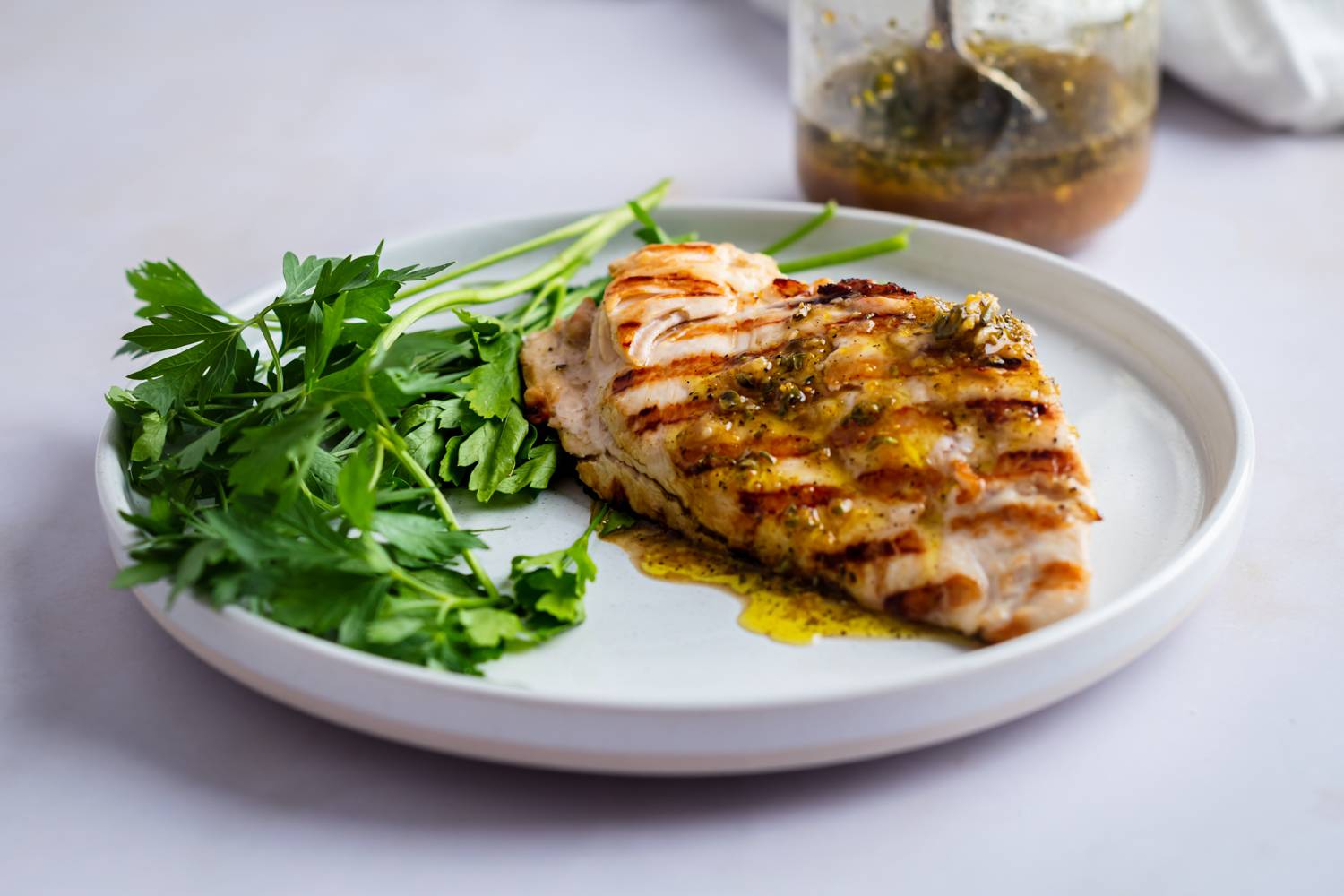 Grilled chicken breast drizzled with herb sauce on a white plate, garnished with fresh parsley. A jar of dressing is partially visible in the background.