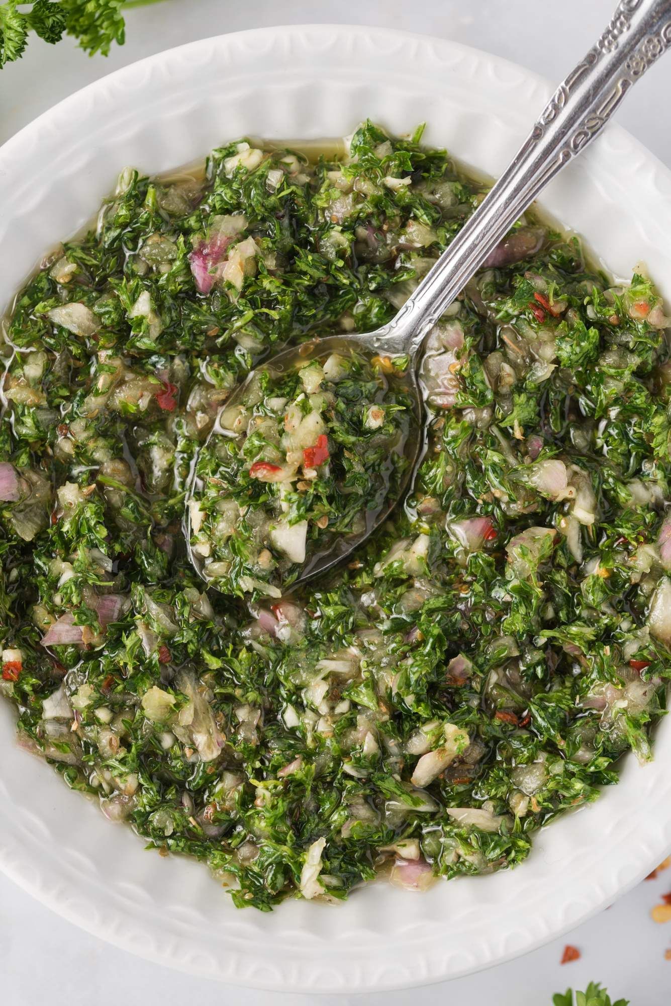 A white bowl filled with homemade chimichurri sauce including parsley, shallots, olive oil, vinegar, red pepper flakes, and salt.